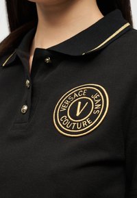 Κοντινό πλάνο μαύρου πόλο πουκάμισου με γιακά με χρυσό περίγραμμα και χρυσό κεντημένο λογότυπο Versace Jeans Couture στο στήθος.