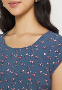 Vestido de patrón floral azul con mangas cortas, que presenta pequeñas flores rojas y blancas sobre una tela texturizada. Escote redondeado, diseño ceñido.