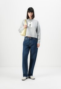 Felpa grigia corta con stampa grafica, jeans larghi blu e scarpe crema. La modella tiene una borsa gialla pallido. Sfondo semplice.