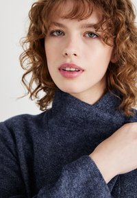 Navy sweater with a high collar, textured fabric, and loose fit, featuring a folded neckline and wide sleeves.