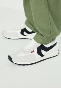 Weiße und graue Sneakers mit schwarzen Akzenten, aus Wildleder und Mesh-Material, flacher Gummisohle und weißen Schnürsenkeln. Kleines rotes Logodetail sichtbar.