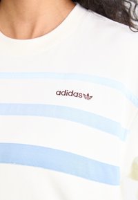 White sweatshirt with horizontal light blue stripes. Features a brown Adidas logo at the chest. Material appears soft with a textured finish.