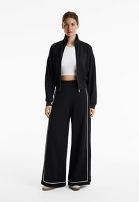 Schwarze Sportjacke mit hochgeschlossenem Kragen und Reißverschluss, kombiniert mit lockeren schwarzen Hosen mit weißen Seitenstreifen und hellfarbigem Schuhwerk.