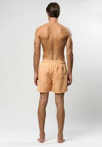 Perzikkleurige zwembroek met een elastische tailleband, achterkantzak en een gladde textuur, van achteraf getoond, gedragen door een shirtloze mannelijke model.