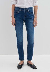 Person i åtsittande blå jeans, vit skjorta med uppkavlade ärmar och svarta spetsiga ballerinas, står mot en enkel ljus bakgrund.