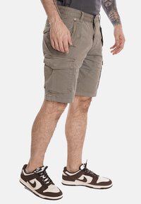 Pantaloni cargo kaki con molte tasche e vestibilità rilassata. Realizzati in tessuto resistente con una texture liscia. Indossati con sneakers marroni e bianche.