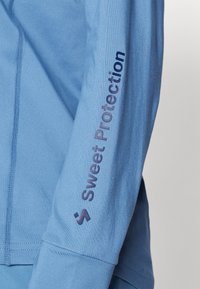 Šviesiai mėlynas ilgomis rankovėmis marškinėliai su glotnia tekstūra. Ant rankovės yra tamsiai mėlyna „Sweet Protection“ logo, o dizainas yra pritaikytas.