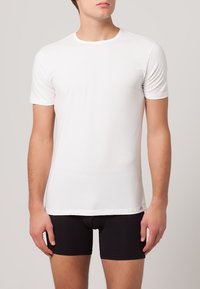 T-shirt bianca in cotone con maniche corte, colletto rotondo e una texture uniforme, indossata sopra pantaloni corti neri aderenti.
