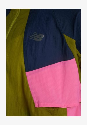 Section de veste bicolore en vert olive, bleu marine et rose avec un logo réfléchissant New Balance sur le tissu bleu marine.