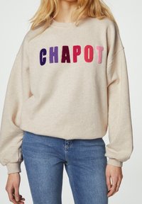 Felpa beige con vestibilità over-size, caratterizzata dalla scritta "CHAPOT" in un ricamo vivace e audace. Maniche lunghe, polsini a coste e un morbido tessuto.