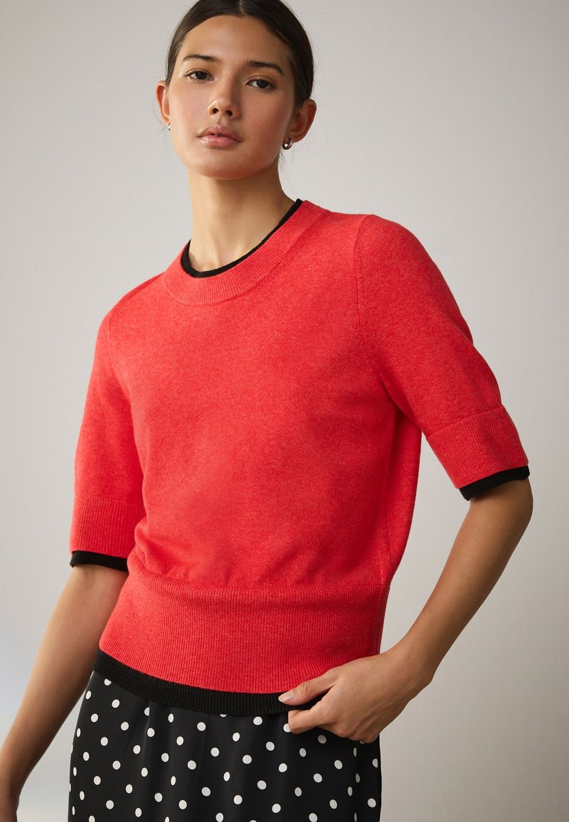 Femme portant un pull rouge vif à manches courtes avec une bordure noire, associée à une jupe noire à pois blancs, debout devant un fond neutre.