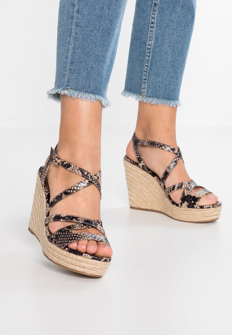 Pieds portant des sandales compensées à lanières à imprimé serpent avec des talons plateforme en jute, associées à un jean bleu délavé à ourlet effiloché.