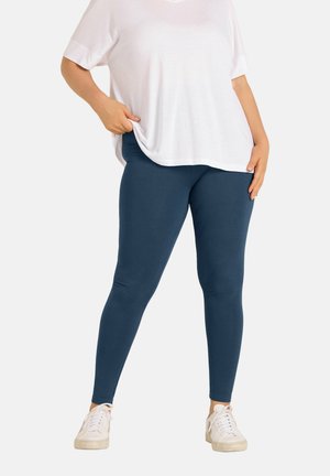 Persoon die donkerblauwe leggings draagt, een los wit T-shirt met korte mouwen en witte sneakers, staand tegen een effen achtergrond.