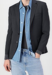 Dunkelgrauer Blazer über einem hellblauen Jeanshemd, kombiniert mit einem schwarzen Oberteil und hellblauen Jeans. Merkmale sind Knöpfe, Taschen und unterschiedliche Texturen.
