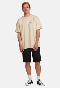 Beige cotton t-shirt with short sleeves and a left chest logo, paired with black shorts and black sneakers, featuring white crew socks.