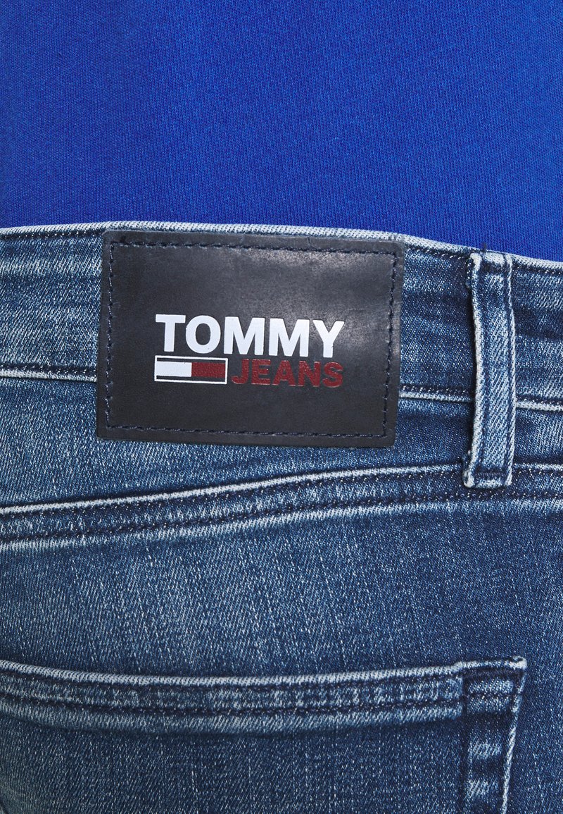 Niebieskie dżinsy z czarną skórzaną metką w pasie, na której widnieje napis "TOMMY" na biało i "JEANS" na czerwono. Widoczne przeszycia i kieszenie.