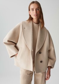 Vrouw in een oversized beige wollen jas met grote revers, zwarte knopen en een bijpassende beige trui en broek, staand tegen een vlakke achtergrond.