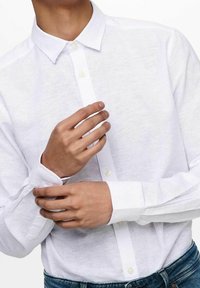 Camisa blanca de lino con cuello clásico y mangas largas. Presenta una textura sutil y botones a lo largo de la parte delantera.