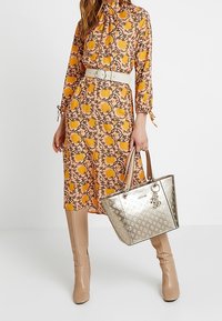 Zlatá metalická taška přes rameno s logem a detaily na přívěsku, kombinovaná s květinovými šaty s páskem v odstínech žluté a broskvové a béžovými botami.
