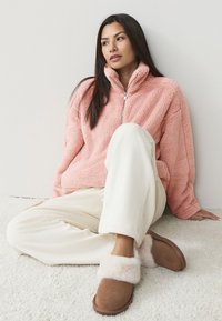 Pile rosa chiaro con colletto alto e zip. Pantaloni in velluto a coste color crema e pantofole marroni con bordo soffice rosa. Texture morbida.