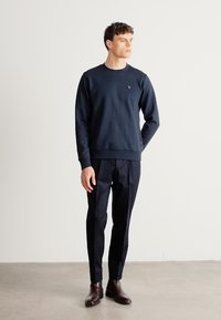 Tmavomodrá mikina s okrúhlym výstrihom a malým logom, skombinovaná s tmavými na mieru šitými nohavicami a hnedými koženými topánkami. Model stojí na svetlom pozadí.