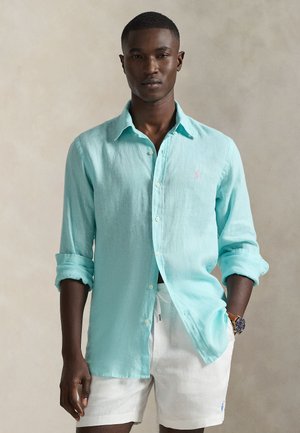 Jasnoniebieska lniana koszula na guziki z podwiniętymi rękawami, z małym różowym logo na piersi, w połączeniu z białymi szortami.