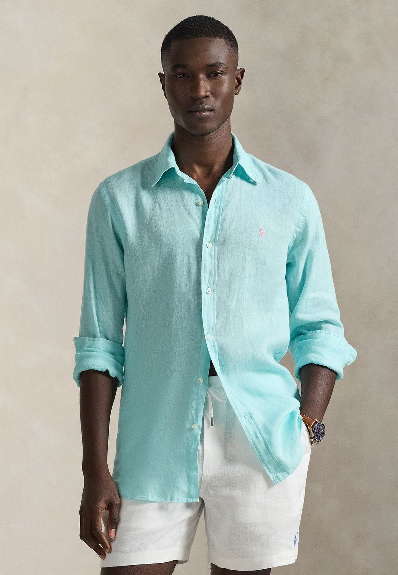 Ανοιχτό μπλε πουκάμισο από λινό με κουμπιά και μανίκια γυρισμένα, με ένα μικρό ροζ λογότυπο στο στήθος, συνδυασμένο με λευκό σορτς.