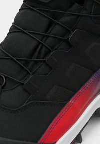 Prim-plan al unei teneși negre cu sfori groase negre, panou lateral texturat și detaliu în degradé roșu deasupra talpă albe.