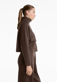 Vrouw in profiel met een bruine cropped jas met hoge kraag en een voorzak, en bijpassende high-waisted broek, haar in een paardenstaart.
