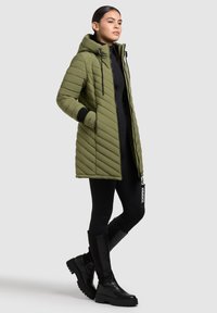 Parka imbottita verde oliva con cappuccio, chiusura frontale con zipper e tasche laterali. Presenta accenti neri e un'etichetta con logo. Indossata con leggings neri e stivali.