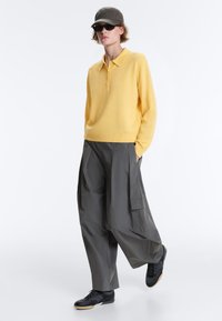 Personne portant un polo jaune à manches longues, un pantalon cargo gris à jambes larges, des baskets noires, des lunettes de soleil noires et une casquette de baseball grise, debout les mains dans les poches.