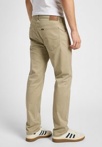 Hombre con pantalones beige de Lee y zapatillas blancas de Adidas, de pie contra un fondo claro liso, visto desde atrás.