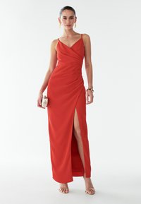 Punane, ilma varrukat pikk kleit, millel on kortsunud disain, sügava külje lõhiku ja siledaga tekstuur. Mudel hoiab väikest metallist kotti ja kannab sandaale.