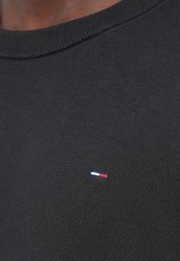 Gros plan d'un pull en maille noir avec un petit logo rectangulaire rouge, blanc et bleu sur la poitrine près du col.