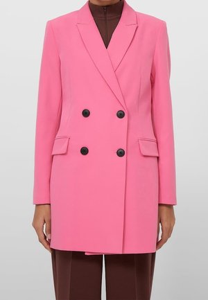 Veste croisée rose vif avec boutons noirs, poches à rabat, portée sur un haut marron zippé et un pantalon marron.