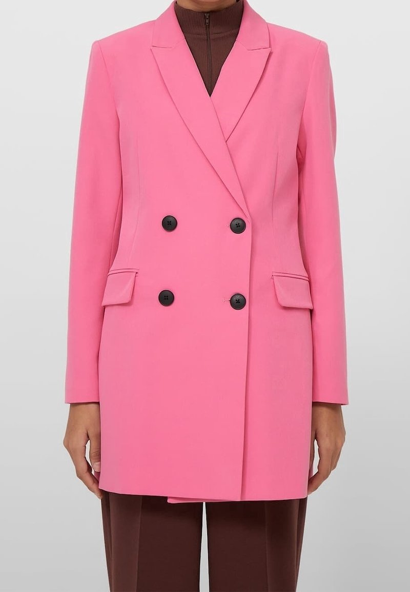 Veste croisée rose vif avec boutons noirs, poches à rabat, portée sur un haut marron zippé et un pantalon marron.