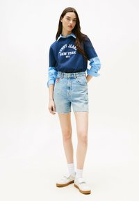 Pantalones cortos de mezclilla en tono claro con el dobladillo deshilachado, combinados con una sudadera azul marino que presenta un logo blanco y una camisa de cuello azul claro debajo. Zapatillas blancas.