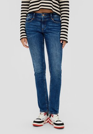 Jean en denim bleu avec une coupe slim, un design à cinq poches et des coutures dorées, associé à un crop top rayé noir et blanc et des baskets.