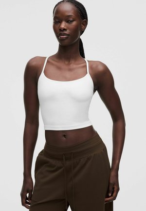 Donna con i capelli intrecciati che indossa un top corto bianco e pantaloni marroni con coulisse, in piedi davanti a uno sfondo chiaro e semplice.
