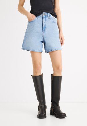 Persona con pantalones cortos de mezclilla azul claro, camiseta sin mangas negra y botas de cuero negras hasta la rodilla, de pie sobre un suelo blanco.