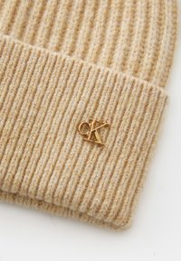 Beige gebreide beanie met ribbelstructuur en een klein gouden "CK"-logo, gemaakt van zacht wolmateriaal en met een nauwsluitende pasvorm.