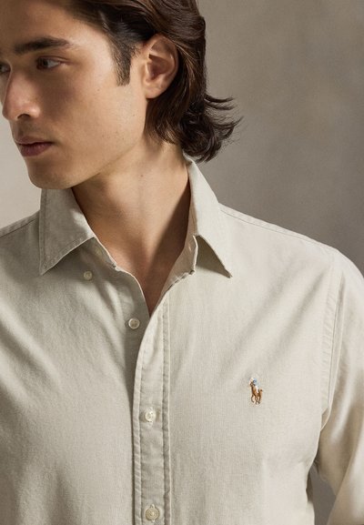 Jonge man met halflang donker haar die een lichtbeige overhemd met knopen draagt, met een klein geborduurd paardenlogo op de borst.