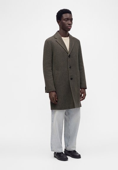 Jeune homme debout portant un manteau marron à simple boutonnage, un jean large délavé clair, des chaussures noires et un haut beige sur un fond uni.