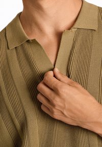 Stickad polo tröja i olivgrön, med vertikal ribbad textur, en klassisk krage och en avslappnad passform. Handjusteringar vid kragen.