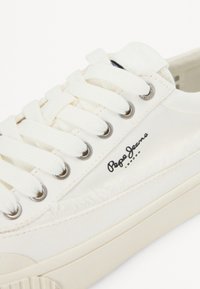 Vita canvas sneakers med texturerade accenter, metallöglor och en svart logotyp. Har flata snören och en gummisula.