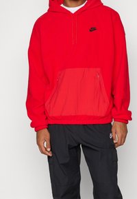 Felpa con cappuccio rosso realizzata in pile, con tasca a marsupio e rifinitura elastica. Logo Nike nero sul petto, con doppia cerniera sulla tasca.