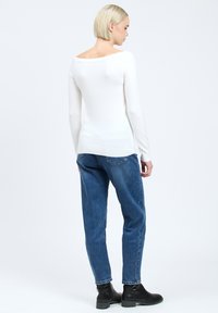 Witte longsleeve top met een brede halslijn, gecombineerd met blauwe high-waisted jeans. Model draagt zwarte enkelboots, gezien vanaf de achterkant.