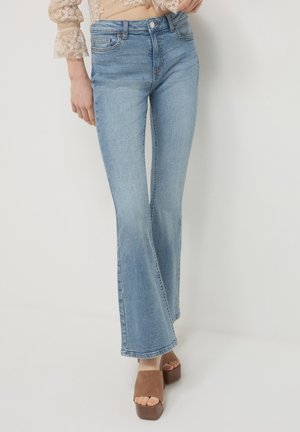 Femme portant un jean flare bleu clair et des sandales à plateforme marron, se tenant contre un fond blanc uni.