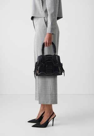 Black handbag with textured leather and intricate hardware details, held by a person in a checkered outfit and black pointed heels.