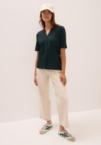 Dunkelgrünes kurzärmeliges Hemd mit Knöpfen, cremefarbene weite Hose und beige Sneakers mit grünen Akzenten. Glatter Stoff und lässige Passform.